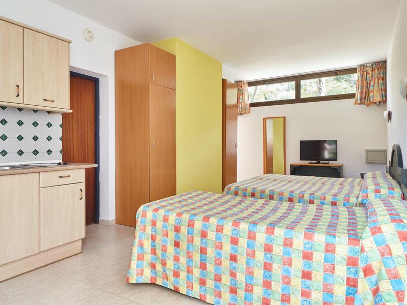 Světle zařízený hotelový pokoj se dvěma jednolůžky, kuchyňským koutem a malou TV.