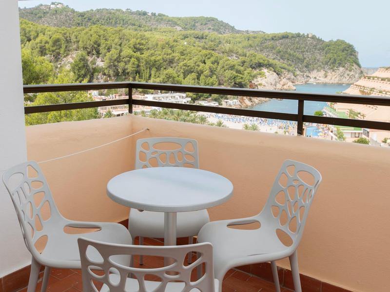 Balkon s kulatým stolem a čtyřmi bílými židlemi s výhledem na zalesněné kopce a moře.