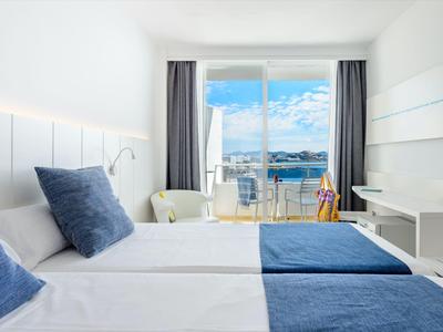 Habitación de hotel luminosa con dos camas y vista a una terraza y el mar.