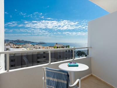 Balcón con mesa y silla blanca, toalla azul, vista al mar y cielo despejado.