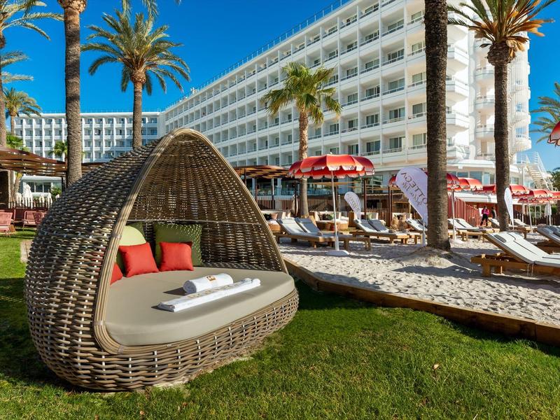 Moderner Liegestuhl aus Rattan vor einem Hotel mit Palmen und Sonnenschirmen