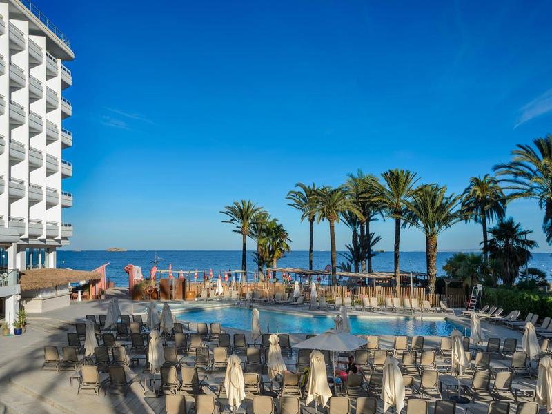 Hotel mit Poolbereich, Sonnenliegen und Palmen am Meer bei klarem Himmel