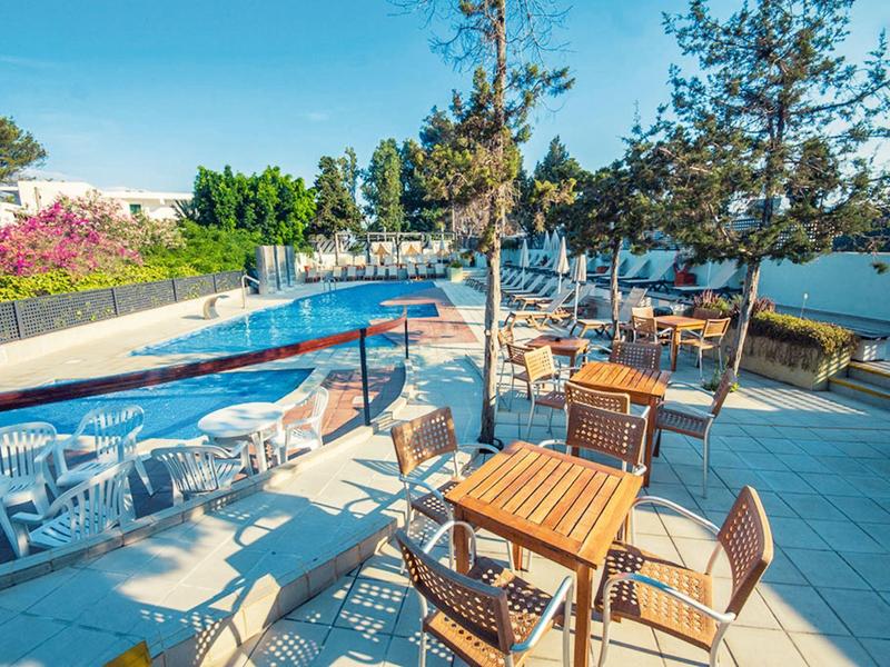 Słoneczny taras hotelowy z drewnianymi i plastikowymi krzesłami wokół basenu z niebieskimi krawędziami.
