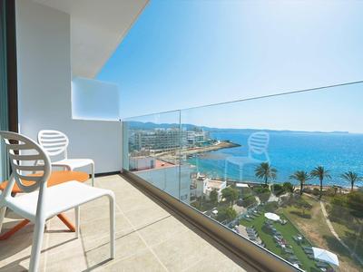 Balkon se židlemi s výhledem na moře, pláž a palmy za jasného nebe.