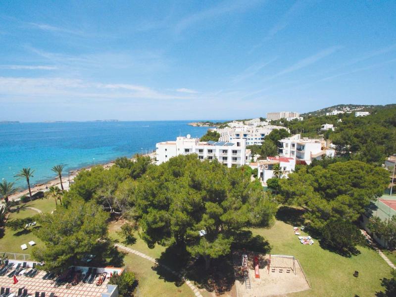 Hotelový komplex s bílými budovami, bujnou zelení a výhledem na modré moře pod jasnou oblohou.