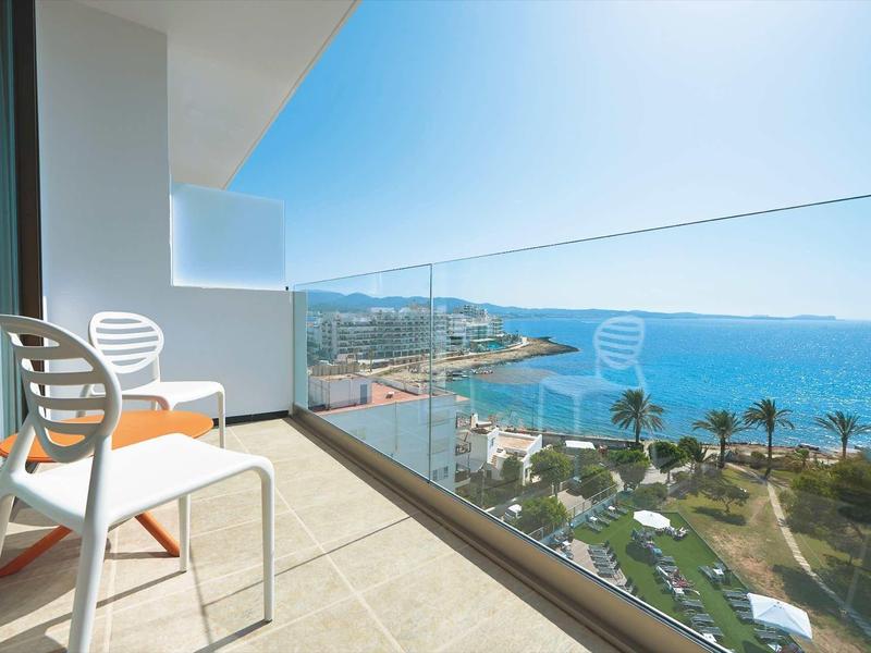 Balkon se židlemi s výhledem na moře, pláž a palmy za jasného nebe.