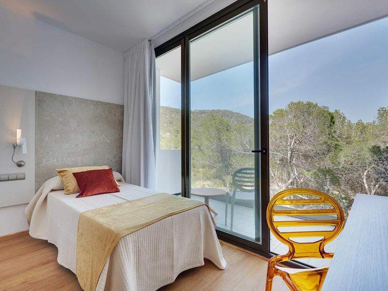 Světý hotelový pokoj s jednolůžkem, skleněnou zdí a výhledem na zalesněné kopce.