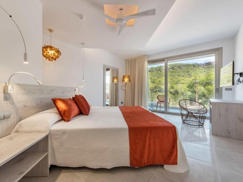 Moderní hotelový pokoj s manželskou postelí, oranžovým přehozem a přístupem na terasu s výhledem do přírody.