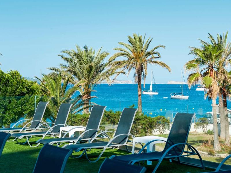 Prázdná lehátka před palmami s výhledem na moře a plachetnicemi v pozadí.