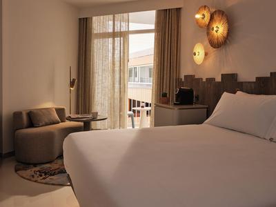 Modernes Hotelzimmer mit großem Bett, Sitzbereich und Balkon mit Blick auf Stadtgebäude.