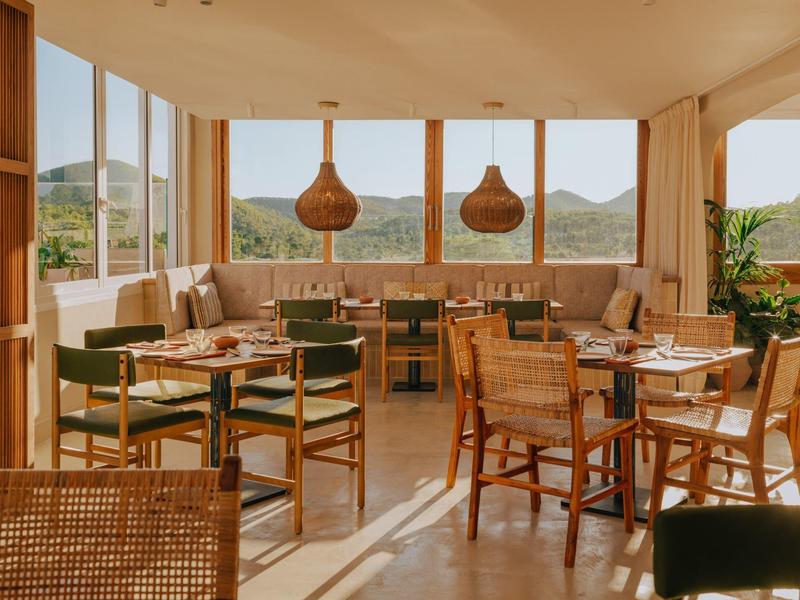 Restaurant lumineux avec grandes fenêtres, mobilier en bois et vue sur des collines verdoyantes.