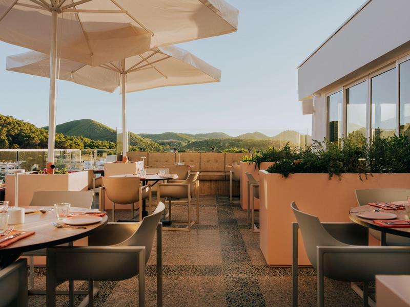 Terrasse d'hôtel ensoleillée avec tables, chaises et vue sur des collines verdoyantes.