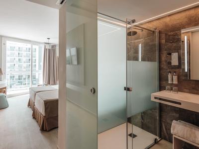 Moderní hotelový pokoj s postelí, relaxačním křeslem a otevřenou koupelnou se skleněnou sprchou.