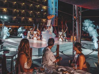Persone sedute a un tavolo guardano una performance di danza all'aperto di sera.