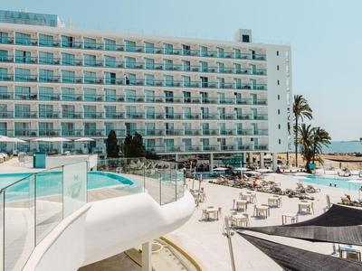 Moderní hotel s bazénem a terasou u moře pod jasnou oblohou.