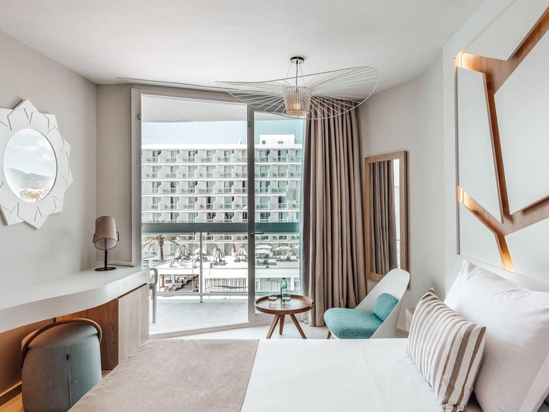 Světle zařízený hotelový pokoj s manželskou postelí, posezením a výhledem na terasu.