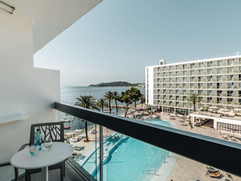 Balkon se stolem a dvěma židlemi s výhledem na bazén, palmy a hotelovou budovu za jasné oblohy.