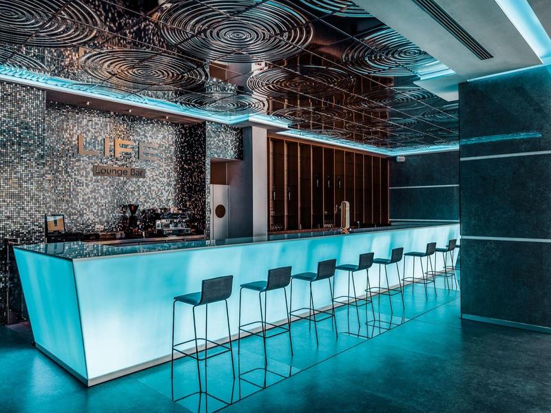 Moderní hotelový bar s osvětleným pultem a barovými židlemi v chladné modré.