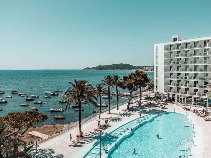 Hotel s velkým bazénem, palmami a výhledem na moře s loděmi a ostrovem v pozadí.