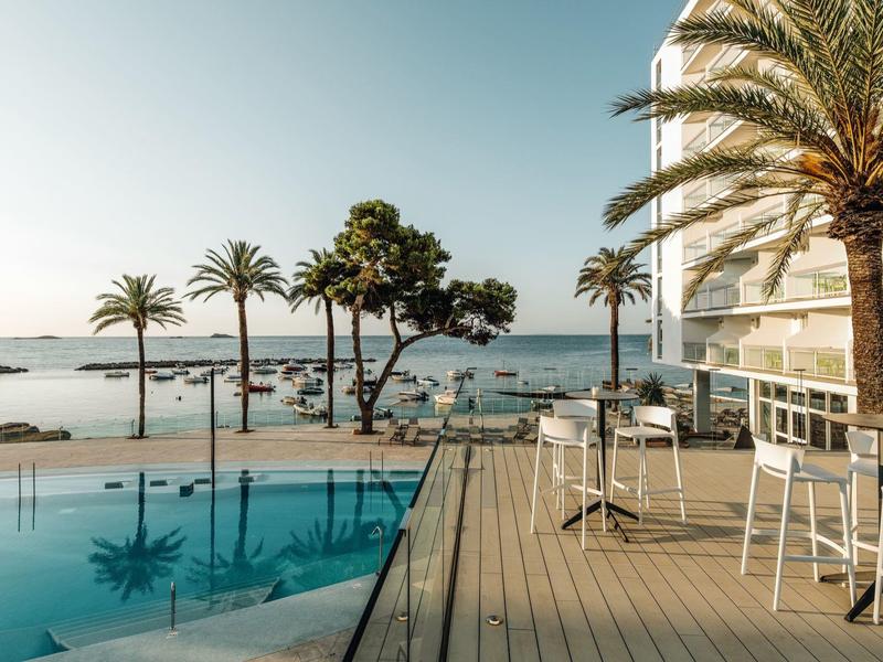 Moderní venkovní bazén s palmami, posezením a výhledem na moře za jasné oblohy.