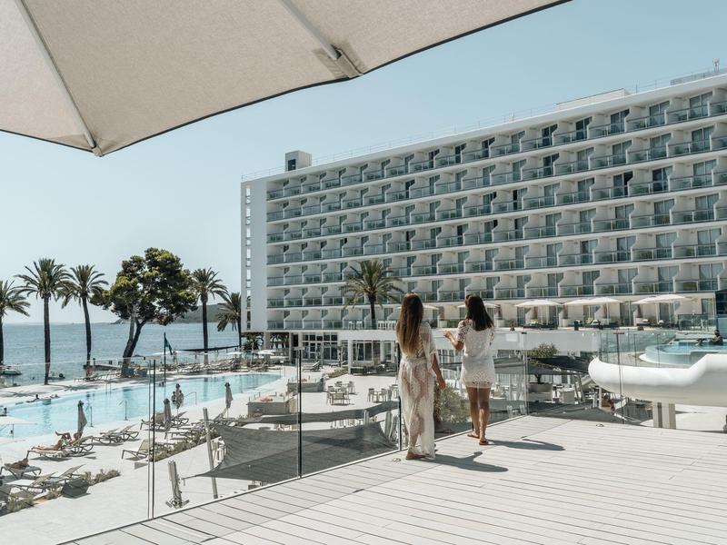 Dvě ženy kráčí u bazénu s lehátky a palmami vedle velkého hotelového objektu u moře.