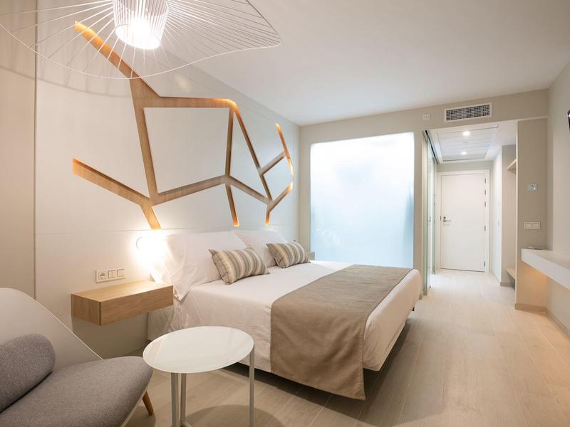 Moderní hotelový pokoj s manželskou postelí, pohovkou, stolem a dekorací na stěně v neutrálních barvách.