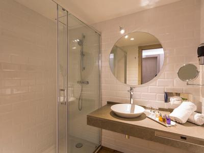 Modern hotel bathroom with glass shower, round mirror, and wooden countertop with amenities.