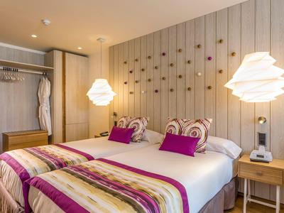 Cozy bedroom with two single beds, purple pillows, striped blankets, wooden wall decor, and bedside table.