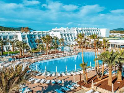 Resort with curved pool surrounded by palm trees, loungers, and white buildings under blue sky.