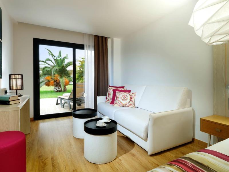 Bright hotel room with white sofa, wooden floor, and garden view through glass door.
