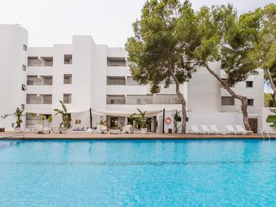 Hotelgebäude mit Pool im Vordergrund und Liegestühlen unter Pinienbäumen.