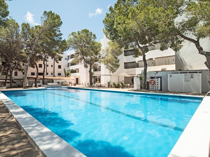 Een helder buitenzwembad omgeven door bomen en witte gebouwen onder een blauwe lucht.