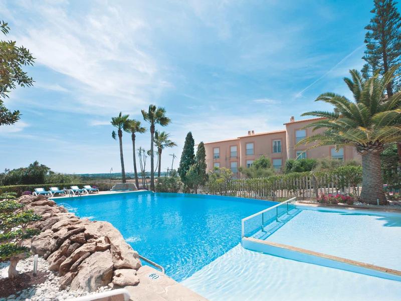 Großer blauer Pool mit Palmen, Steinen und rosa Gebäude unter blauem Himmel bei Tageslicht.
