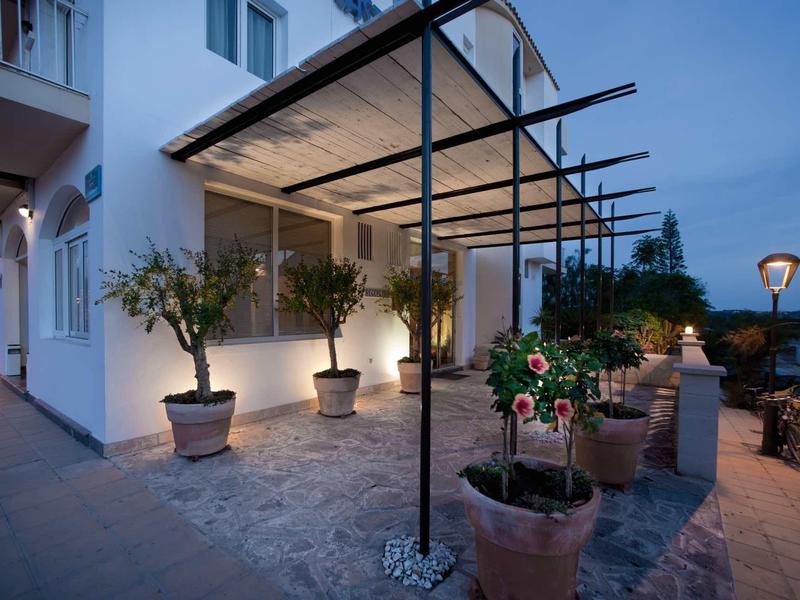 Terrasse mit Pflanzkübeln und Pergola, beleuchtet bei Dämmerung, angrenzend an weißes Haus.