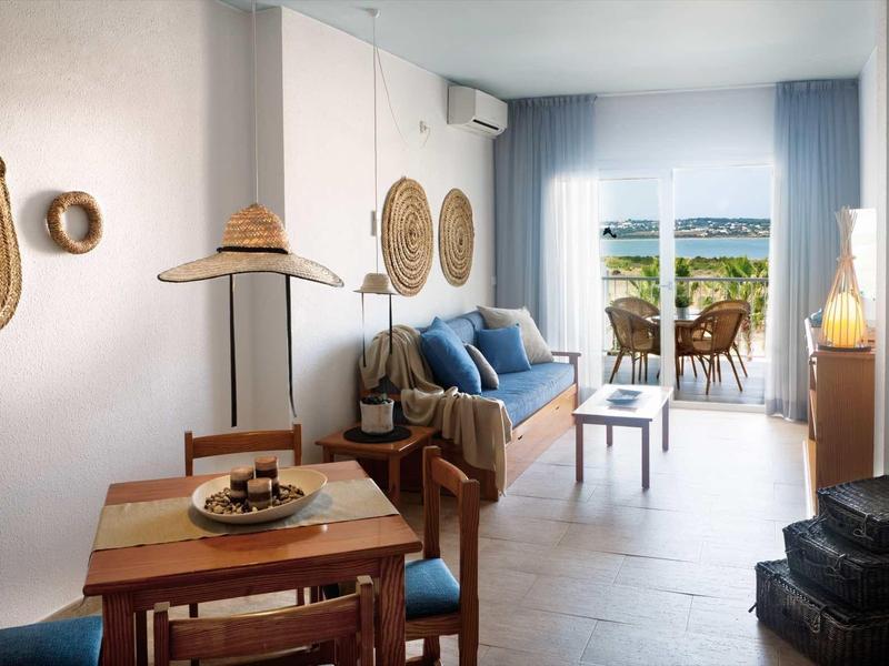 Gemütliches Wohnzimmer mit Holzmöbeln, blauen Kissen, Wanddekoration und Terrassentür mit Ausblick.