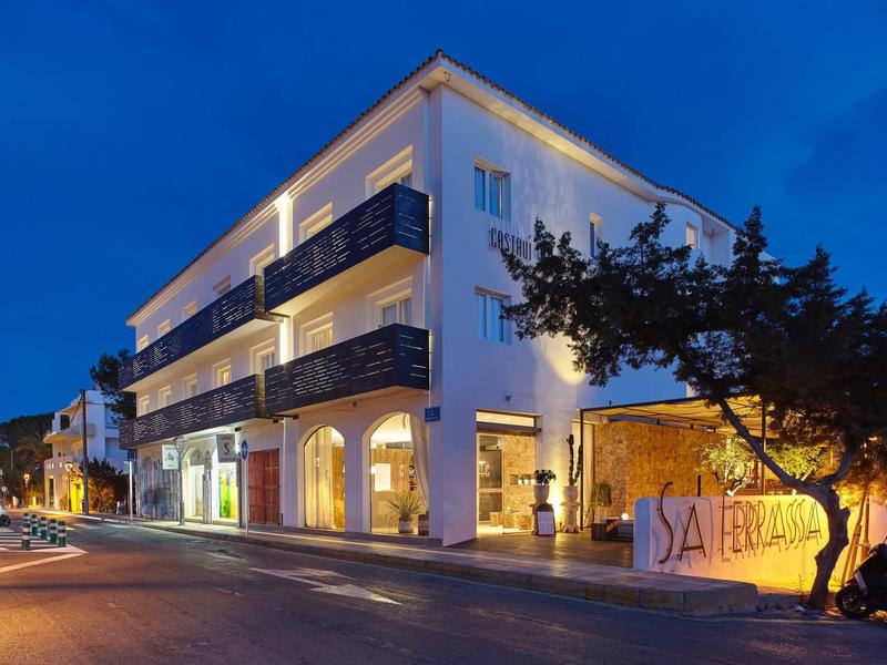 Weißes Gebäude mit Balkonen bei Nacht, beleuchtet, Baum im Vordergrund, ruhige Straße.