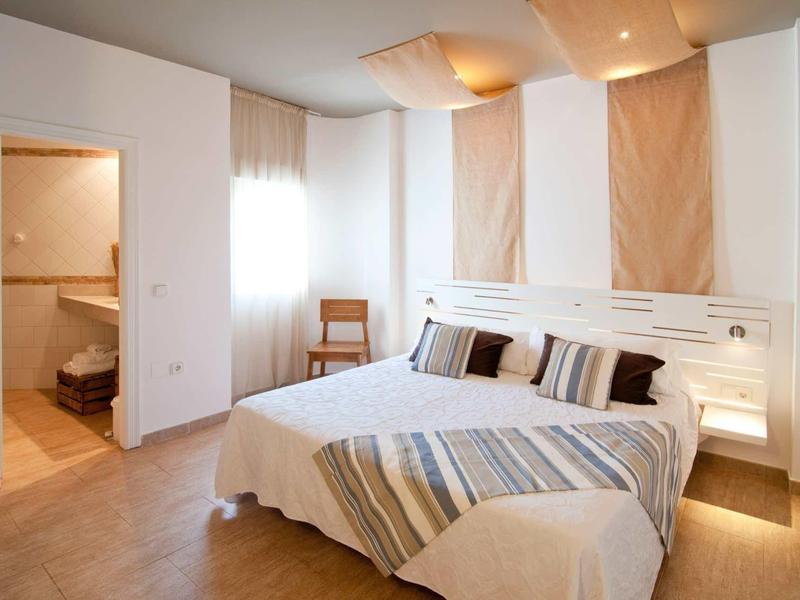 Helles Schlafzimmer mit Doppelbett, gestreiften Kissen, Holzstuhl und angrenzendem Bad.