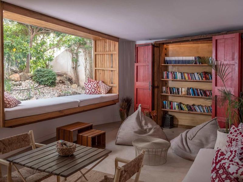 Gemütlicher Leseraum mit großen Fenstern, Holzregal, Sitzsäcken und Pflanzen am Fensterplatz.