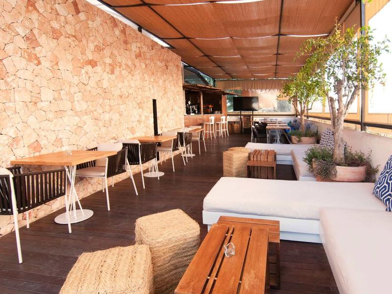 Offene Terrasse mit Holzboden, gepolsterten Sitzbänken und Tischen neben einer Steinwand.