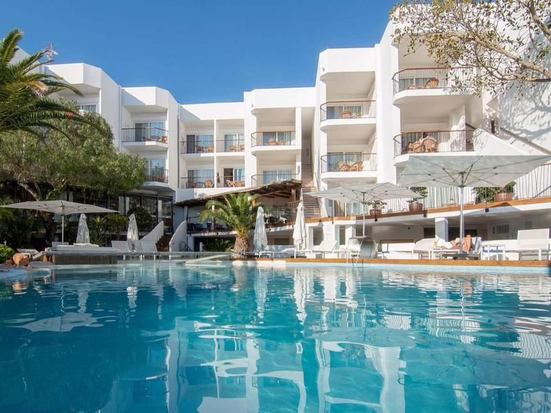 Moderner weißer Hotelkomplex mit Balkonen, großem blauen Pool und Palmen unter klarem Himmel.