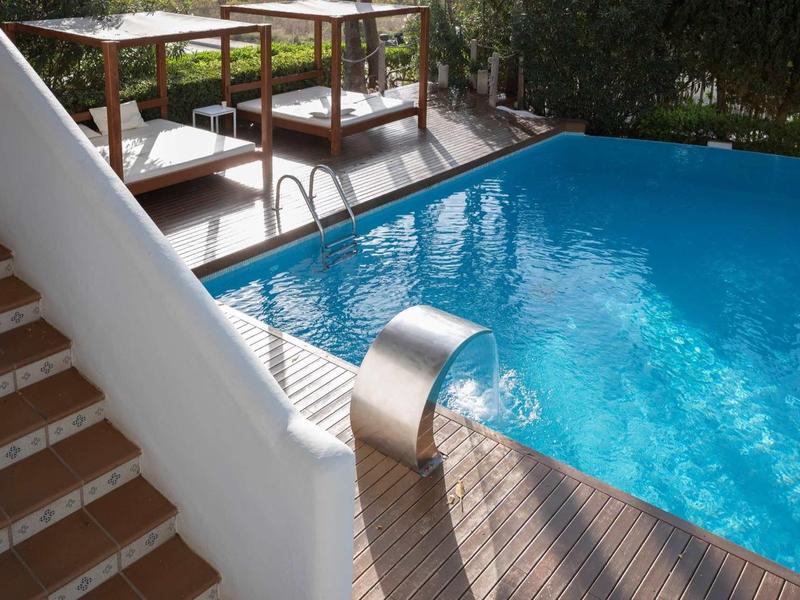 Blauer Pool mit Wasserfall im Garten, neben weißen Treppen und Holzliegen bei Sonnenschein.