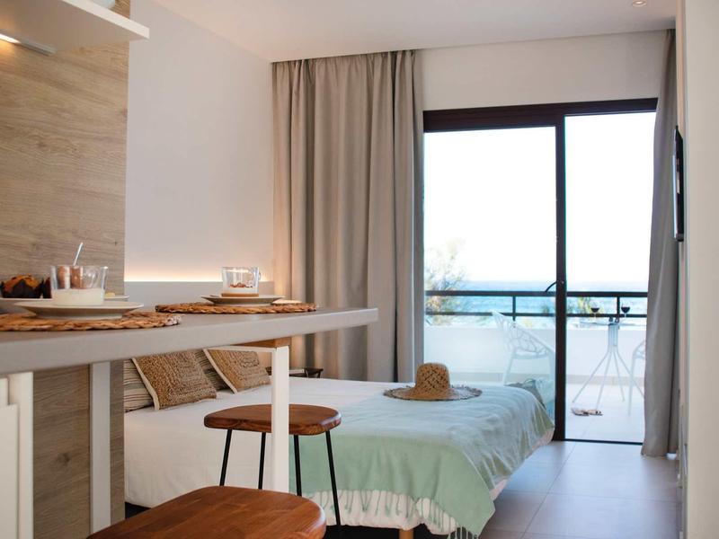 Helles, modernes Schlafzimmer mit Einzelbett, Holzstühlen und großem Fenster mit Blick aufs Meer.