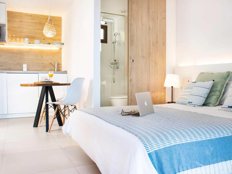 Helles Schlafzimmer mit weißer Bettdecke, Holzwand, kleinem Tisch und moderner Küchenecke.