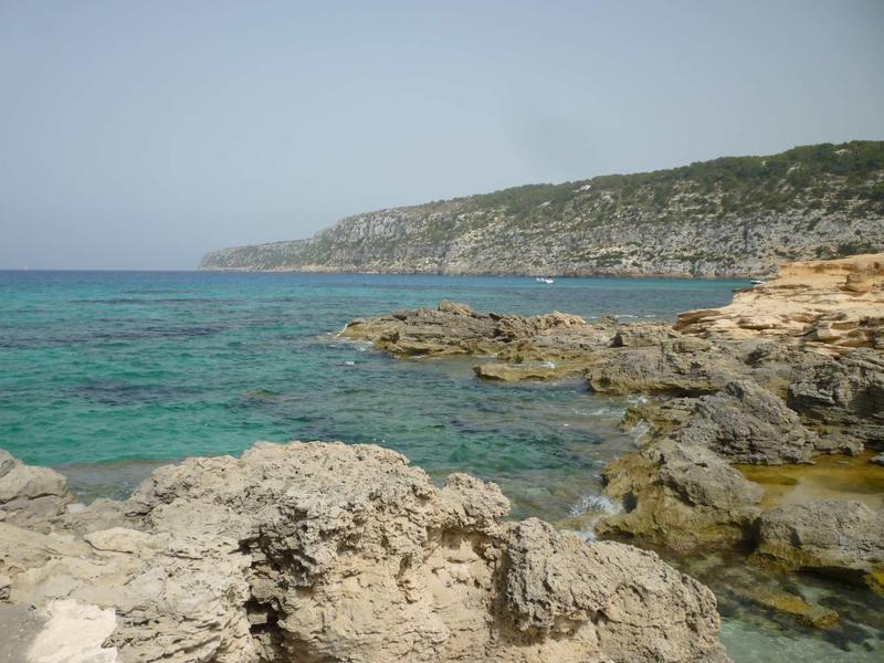 Klares Meer mit türkisfarbenem Wasser, felsige Küste und bewachsene Hügel im Hintergrund.