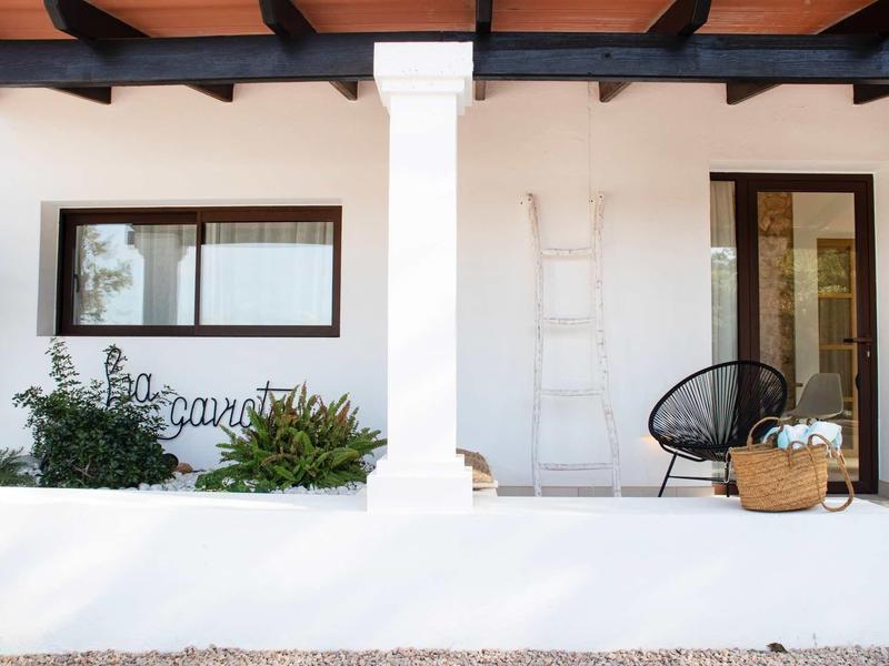 Weißes Haus mit schwarzem Holzgitterdach, zwei Fenster und Sitzplatz mit Korbstuhl und Leiter.