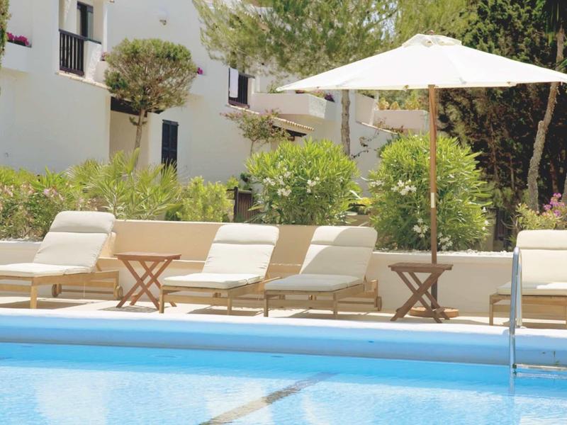 Strefa basenowa z leżakami, parasolem i zielenią przed białymi budynkami.