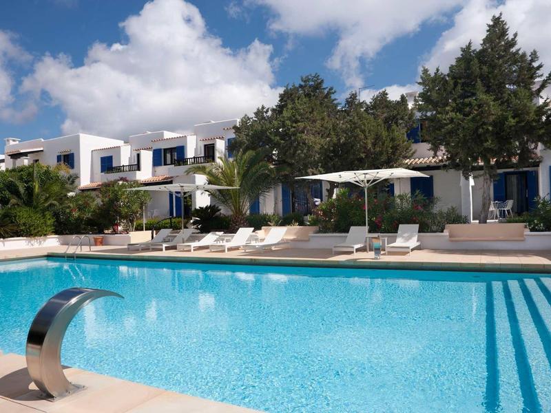 Zapraszający basen hotelowy z leżakami i parasolami przed białym budynkiem pod niebieskim niebem.