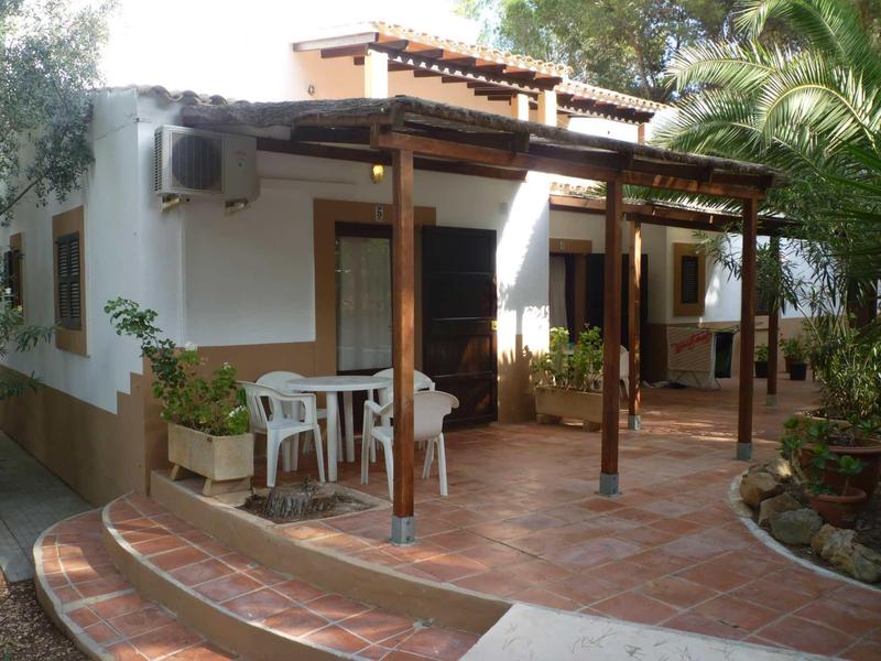 House with covered terrace, white chairs and tables, surrounded by plants.