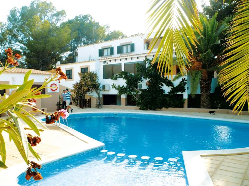 Pool in front of a white house with green shutters, surrounded by trees.
