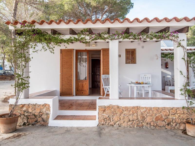Casa de vacaciones blanca con terraza abierta, ventanas de madera y plantas trepadoras en jardín soleado.